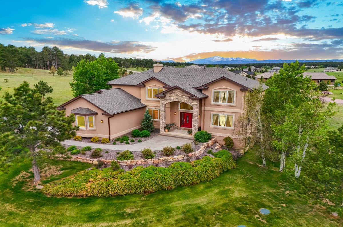 Aerial view of a large two-story residential house with beige stucco exterior, stone accents, and a bright red front door. The property features a manicured front yard, green trees, and a paved driveway, with a natural landscape and snow-capped mountains under a sunset sky in the background.